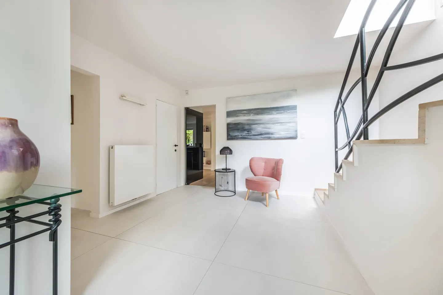 Bright, modern hallway with white walls, a pink chair, and a black metal staircase railing. A painting hangs above the chair.