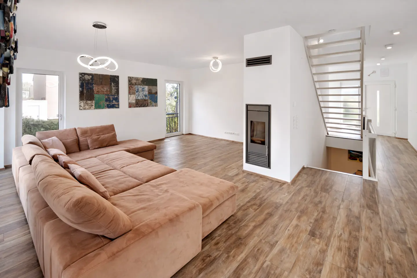 Bright living room with a brown sectional sofa, wood floors, and white walls. A modern fireplace and staircase are visible. Artwork hangs above the sofa.