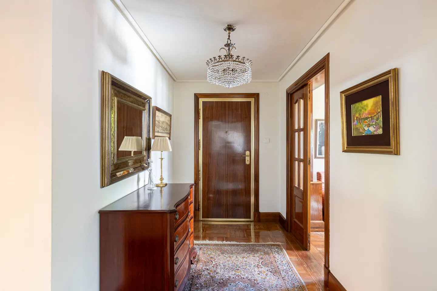 Bright hallway with a wooden dresser, ornate crystal chandelier, patterned rug, and a dark wood door at the end.