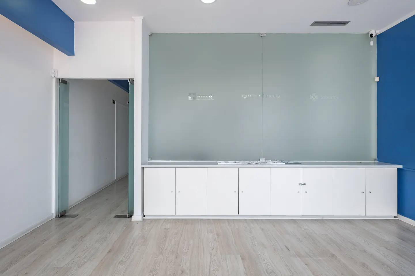 Bright office space with a glass wall, white cabinets, and light wood floors. A glass door leads to another room. Blue accent wall on the right.