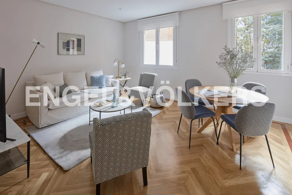 Bright living room with herringbone wood floors, a white sofa, round table, and gray chairs. Windows let in natural light.