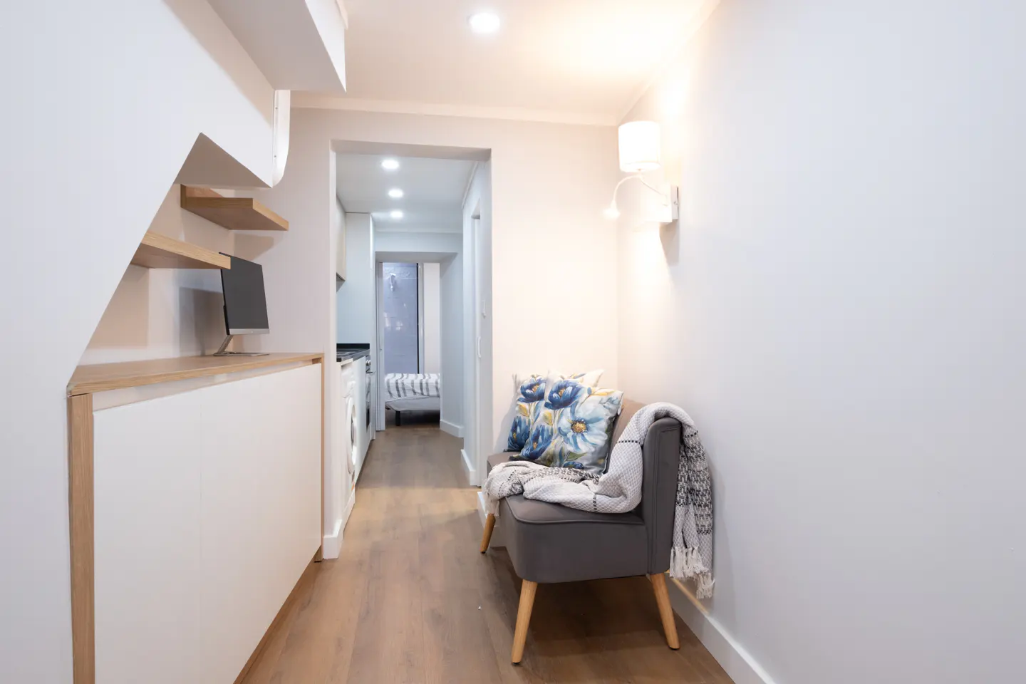 Hallway with a gray chair, blue floral pillows, and a white blanket. A desk with a monitor is under the stairs. A washing machine is in the background.