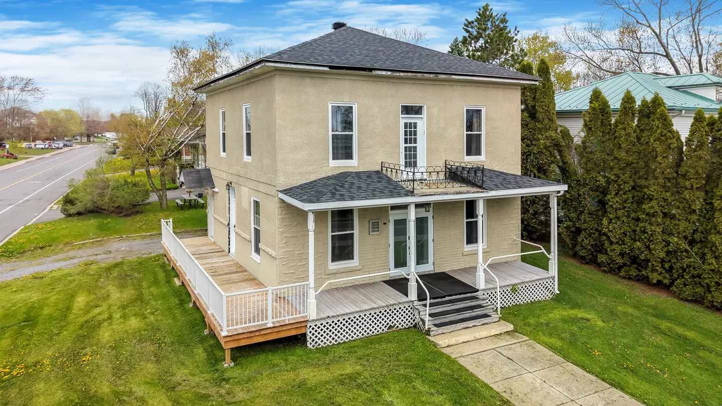 Two-story beige house with a gray roof, white trim, and a porch. Green lawn and trees surround the property.