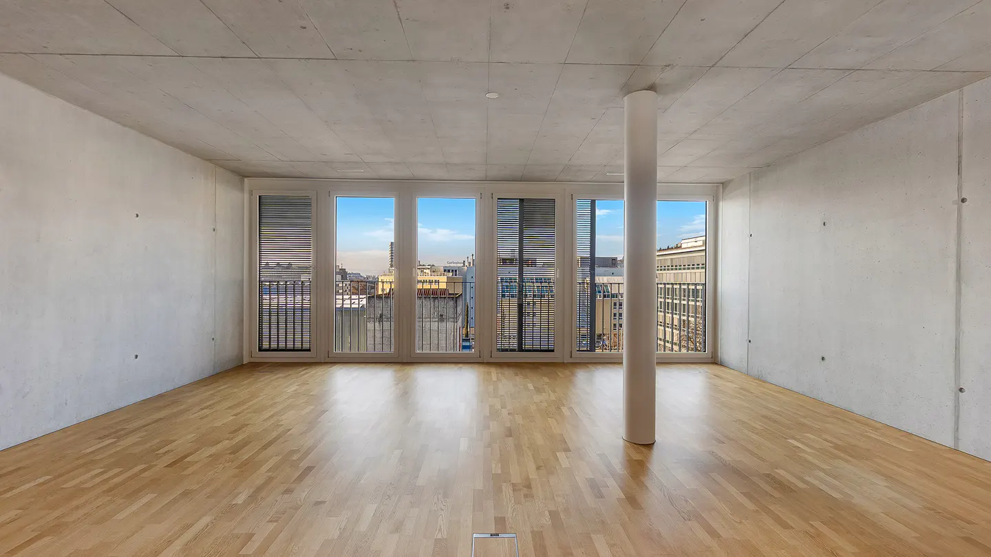 Empty room with hardwood floors, concrete walls, and large windows showing a city view. A white column stands near the windows.