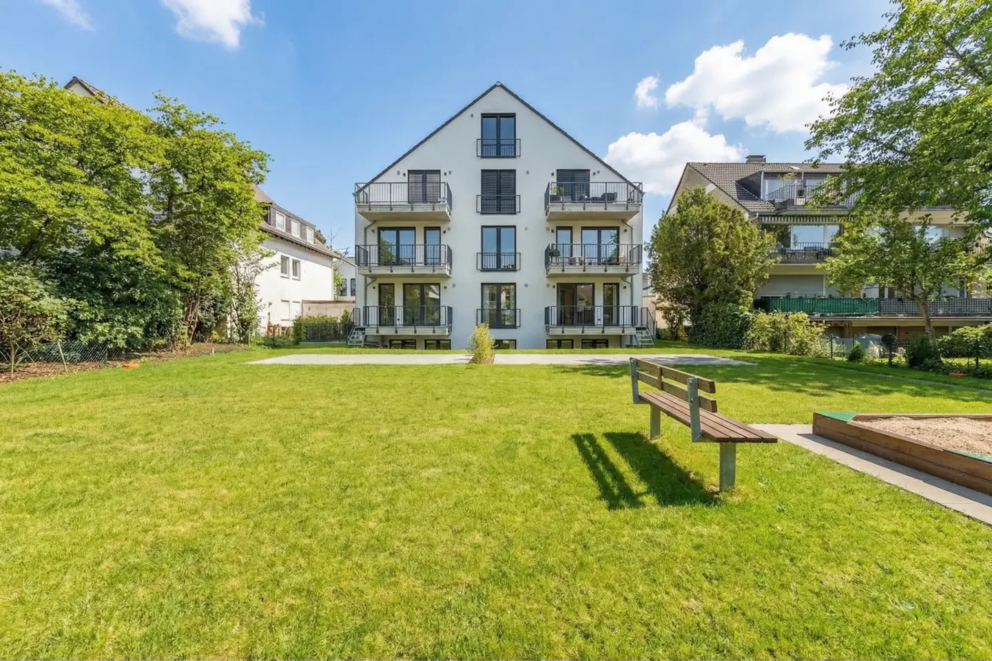 Three-story white apartment building with black balconies, set on a green lawn with a bench and sandbox.