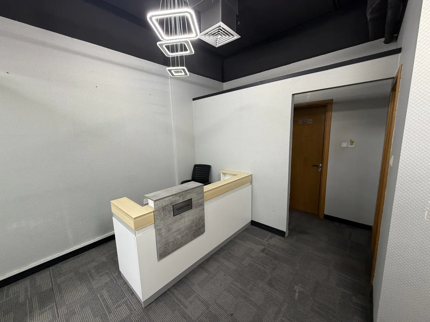 A modern office reception area with a white desk, gray accents, and a black chair. Overhead, square LED lights illuminate the space.