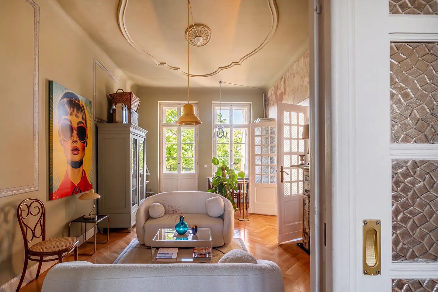 Living room with white sofas, wood floors, and a painting of Audrey Hepburn. French doors lead to another room.