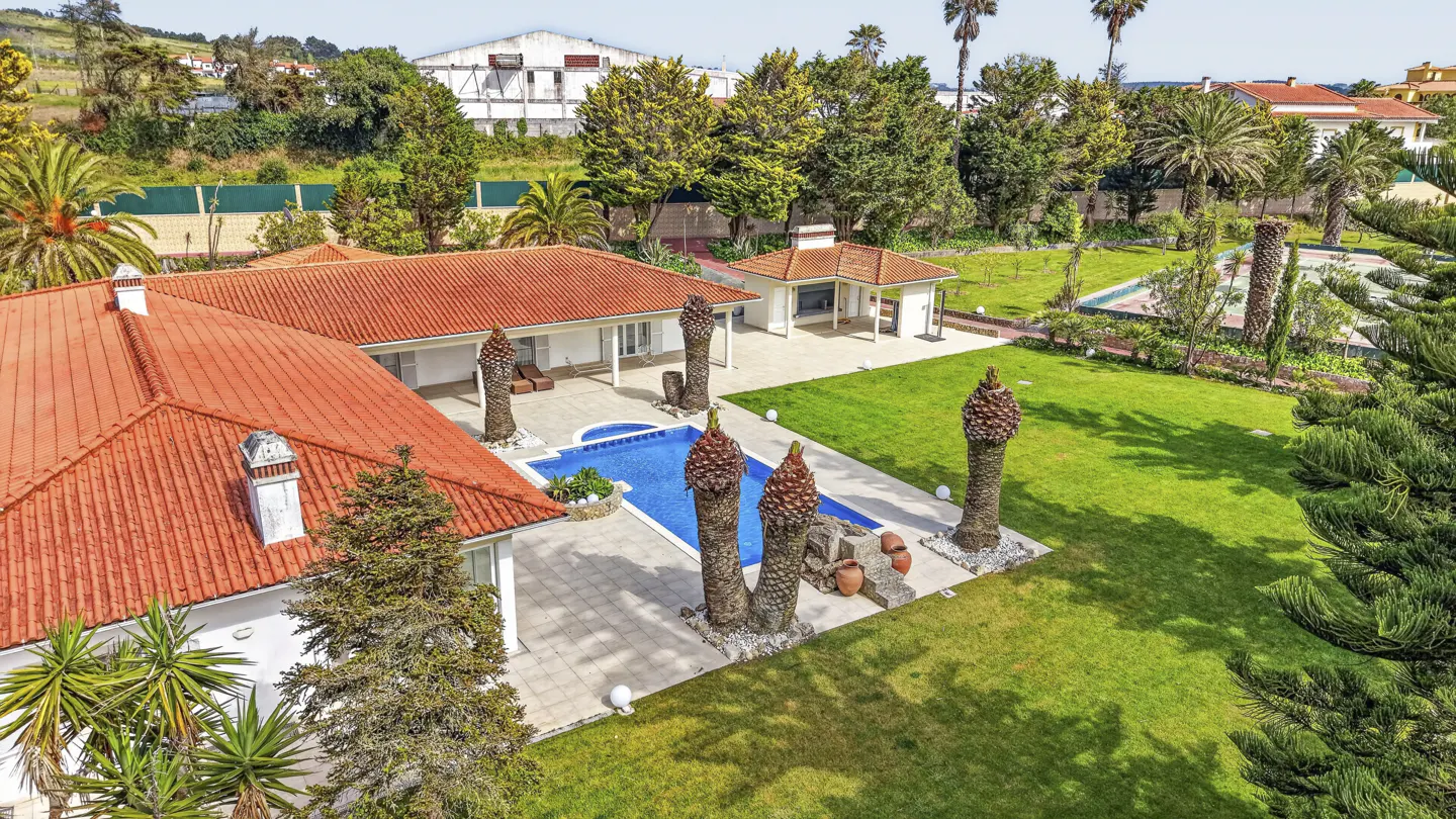 Aerial view of a white house with a red tile roof, a blue swimming pool, and a large green lawn with palm trees.