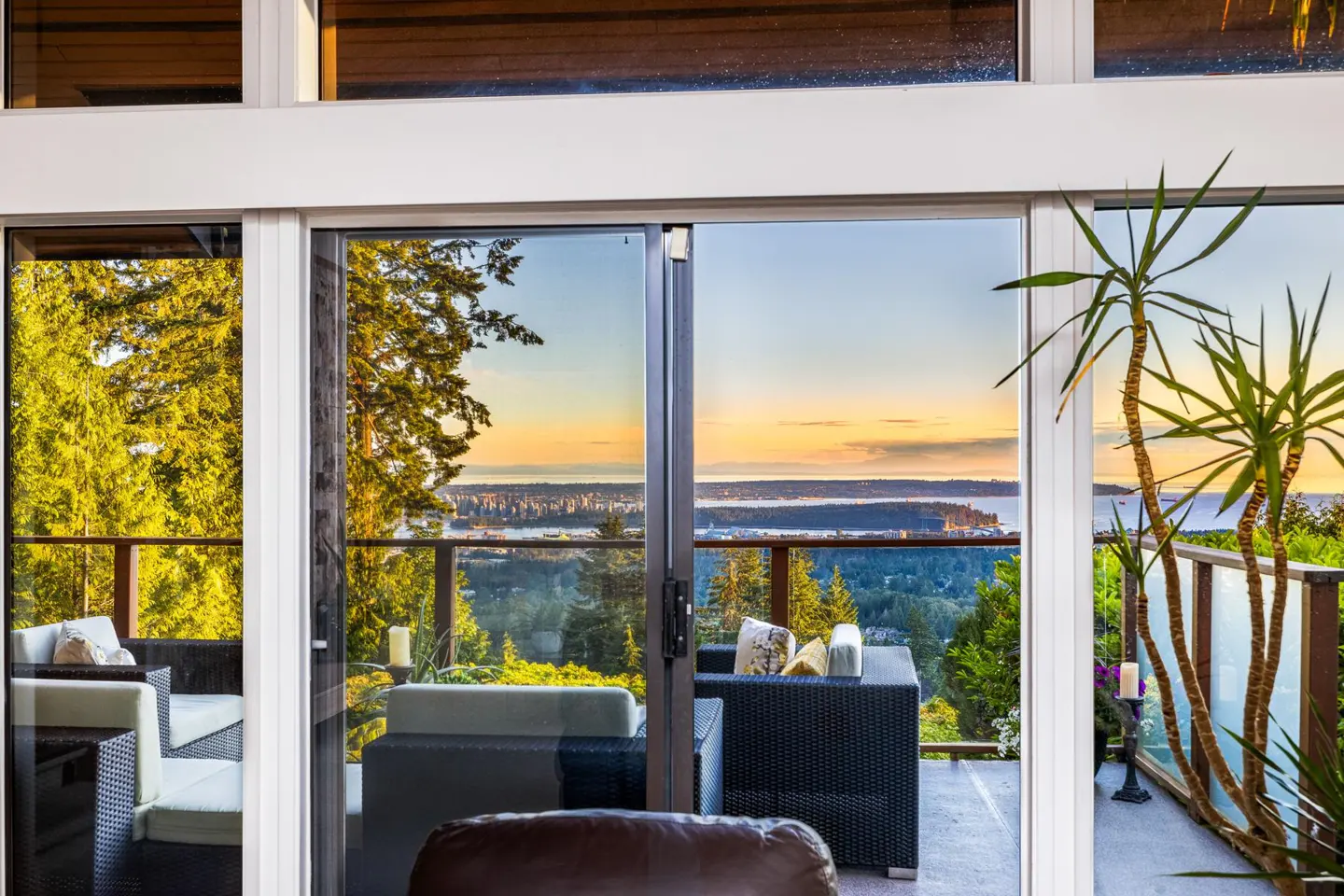 View from a sliding glass door onto a patio with wicker furniture and a scenic view of water and trees at sunset.
