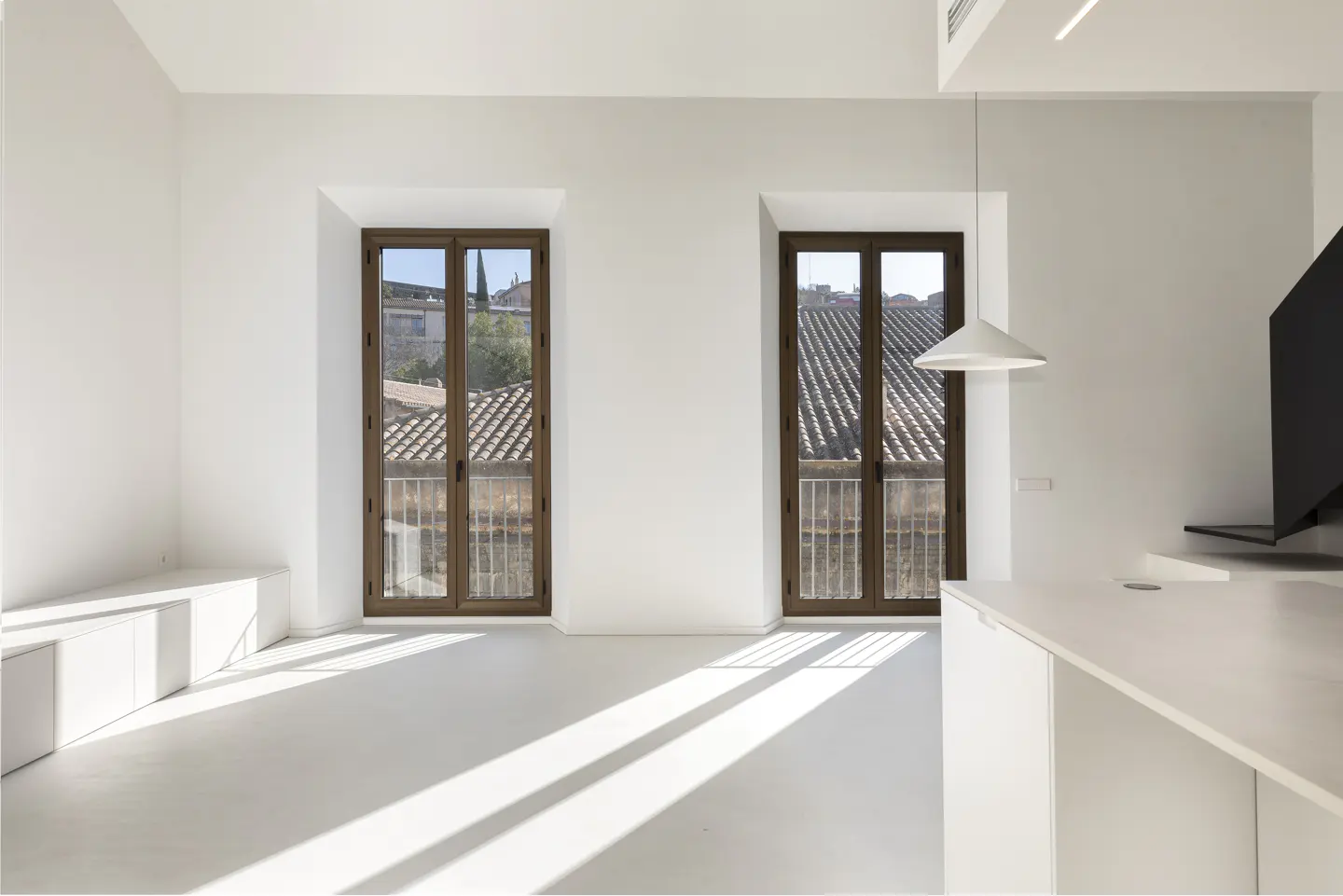 Bright, white room with two brown-framed windows showing rooftops. A white kitchen island and a modern light fixture are visible.
