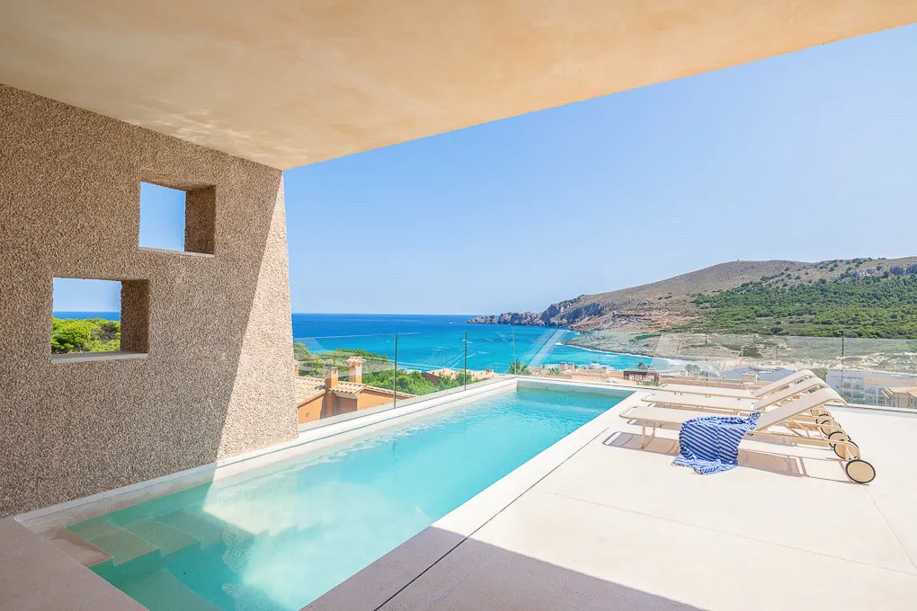 Infinity pool on a terrace with lounge chairs overlooking a turquoise sea, coastal hills, and a clear blue sky.