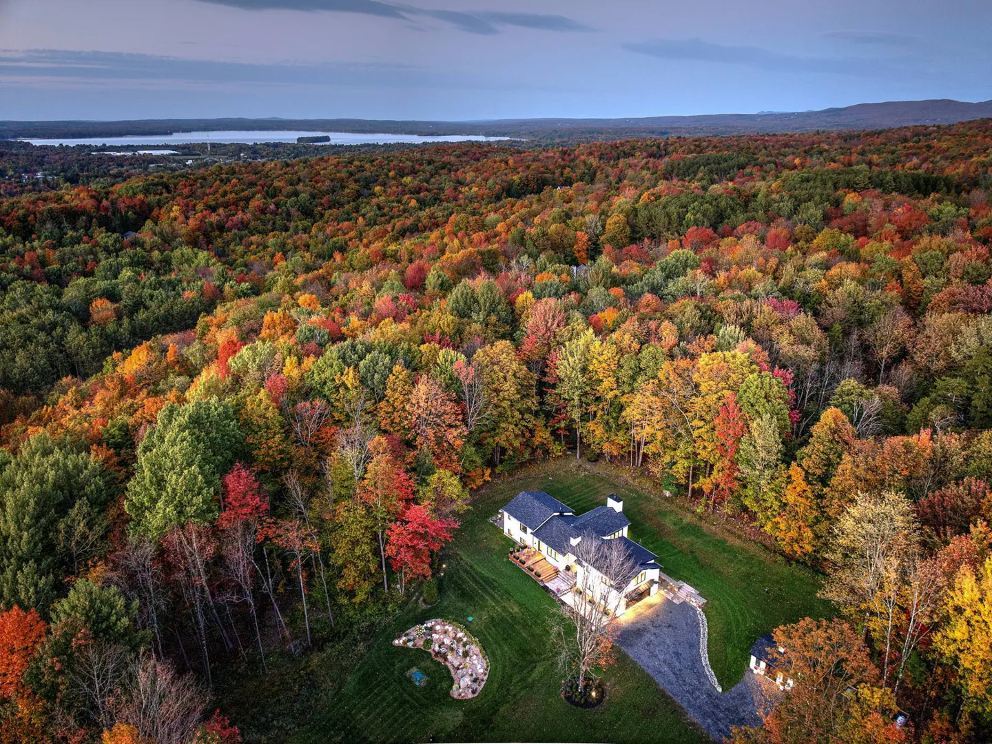 Aerial view of a large, white house with a dark roof surrounded by colorful autumn trees. A lake is visible in the distance.