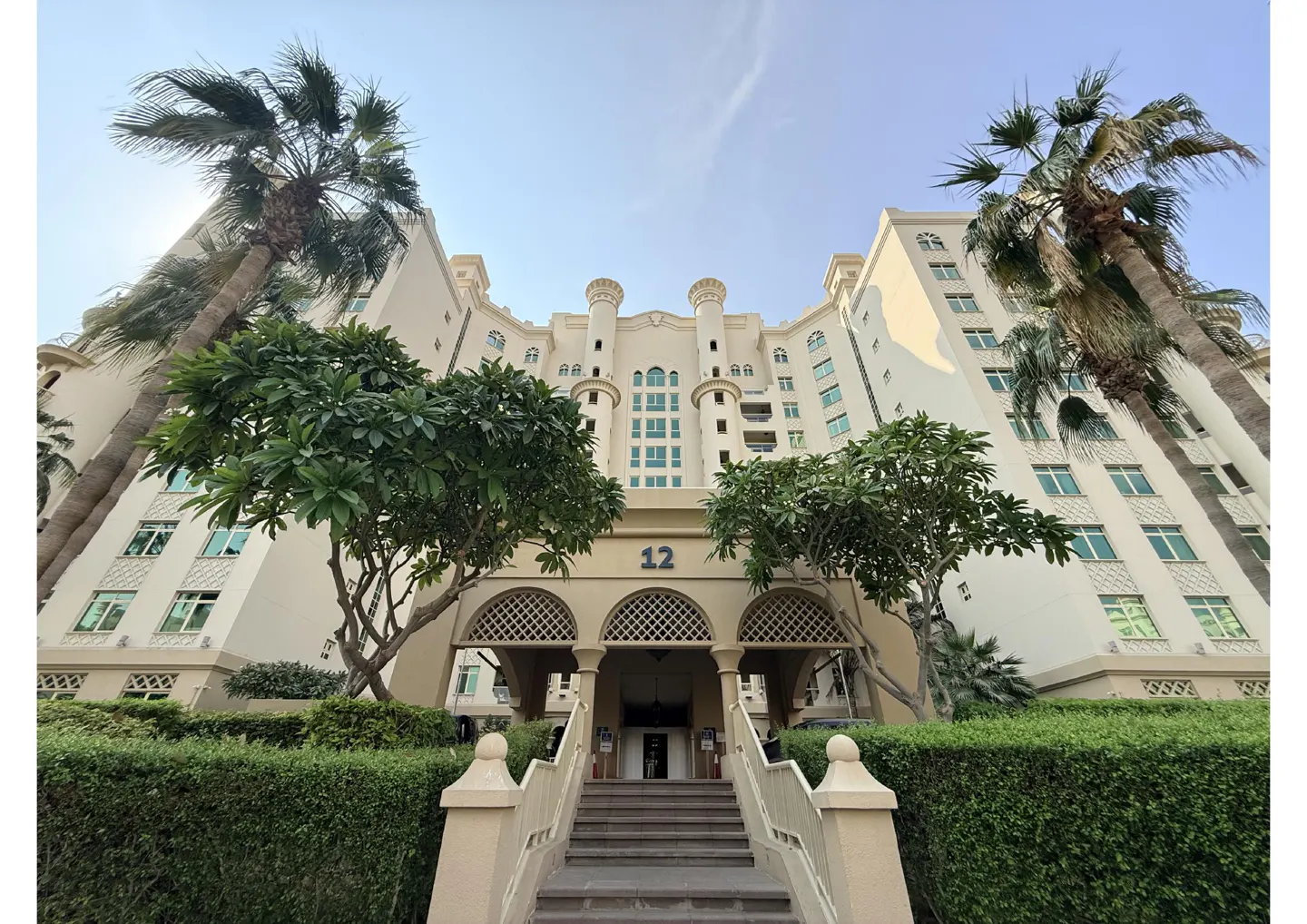 Exterior view of a cream-colored building with palm trees and green bushes. The building has a number 12 above the entrance.