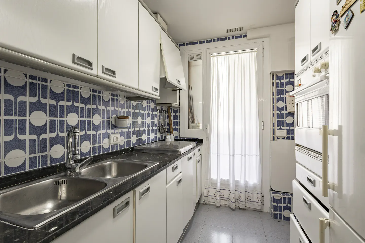 A kitchen with white cabinets, a black countertop, and a stainless steel sink. Blue and white patterned tiles line the wall. A window with white curtains is visible.