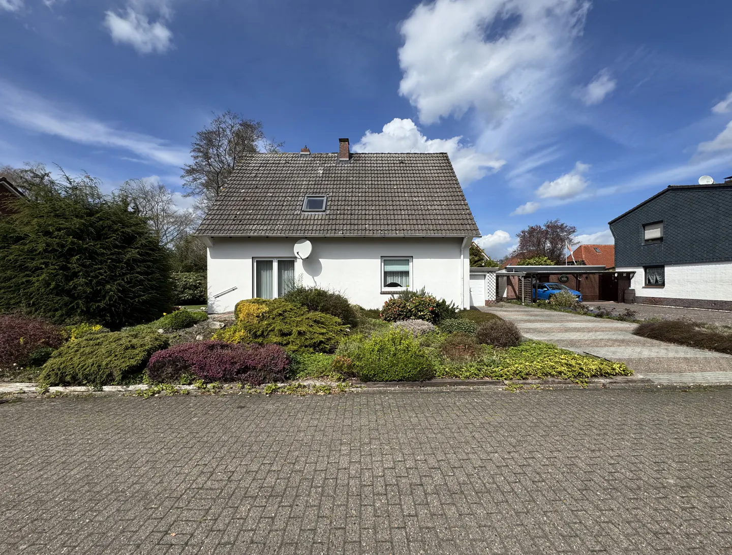 A white, one-story house with a gray roof and a satellite dish, surrounded by colorful bushes and a brick road.