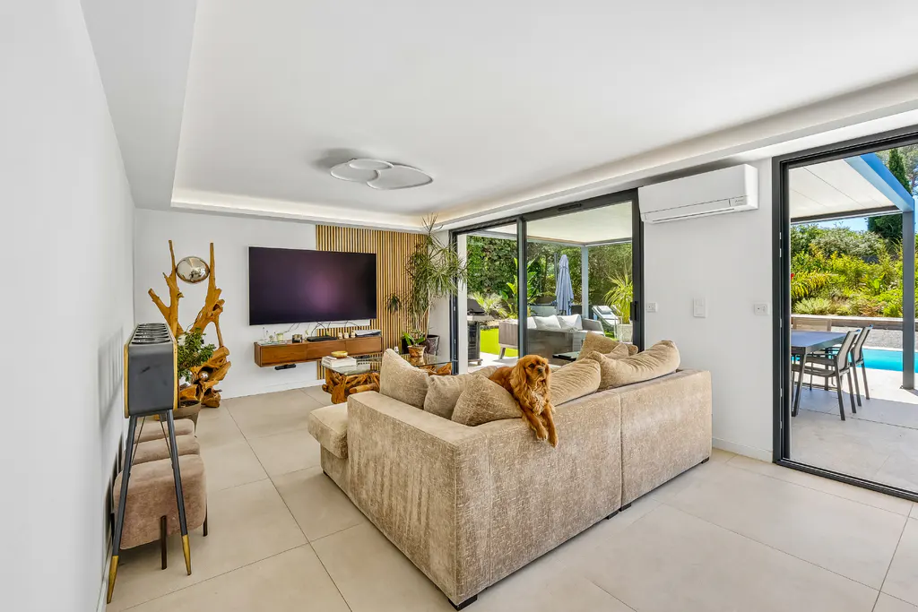 Bright living room with a tan sofa, a dog, and sliding glass doors to a patio with a pool. A large TV is mounted on the wall.