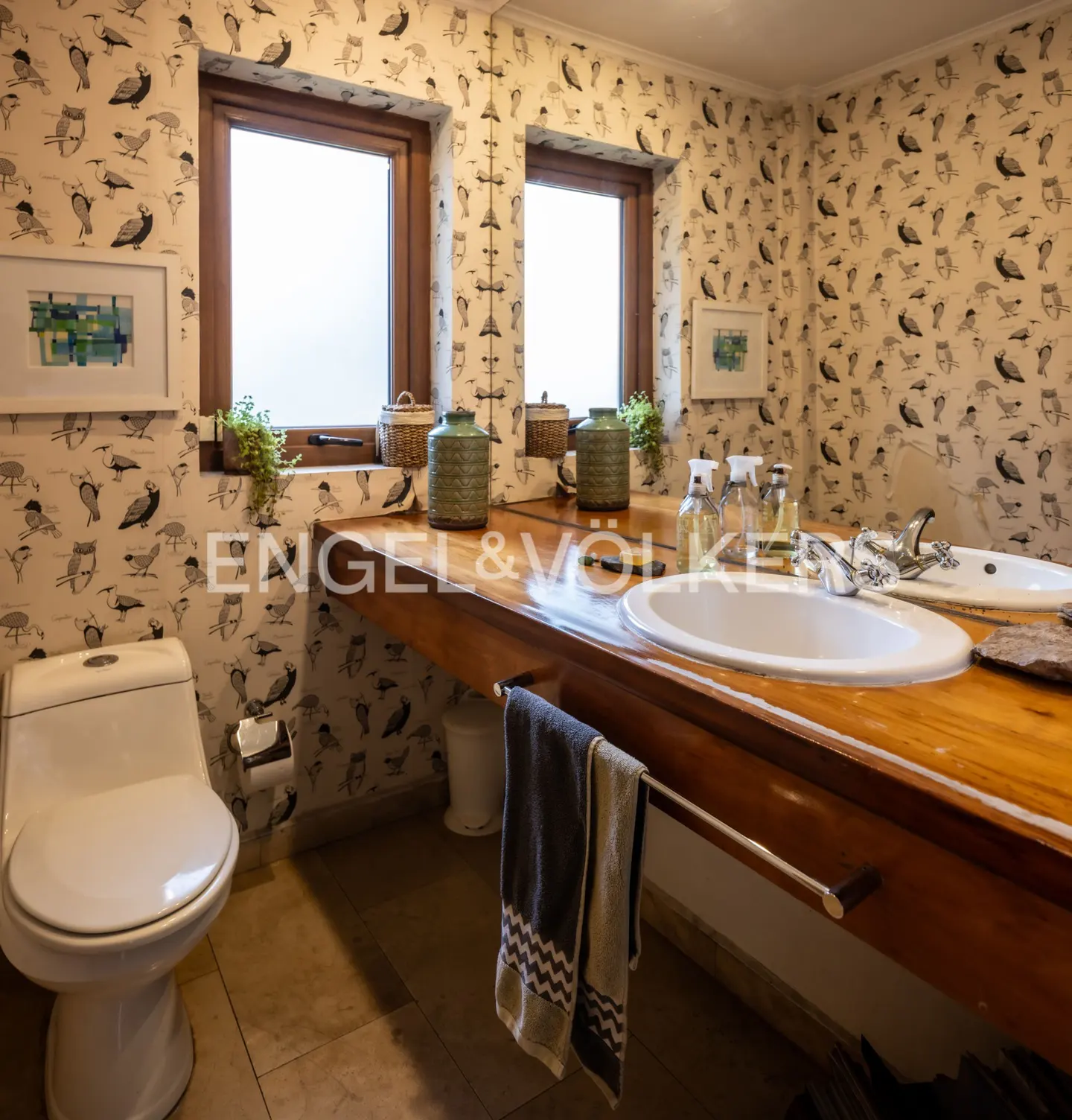 Bathroom with bird-patterned wallpaper, wooden countertop with two sinks, and a white toilet. Two windows provide natural light.