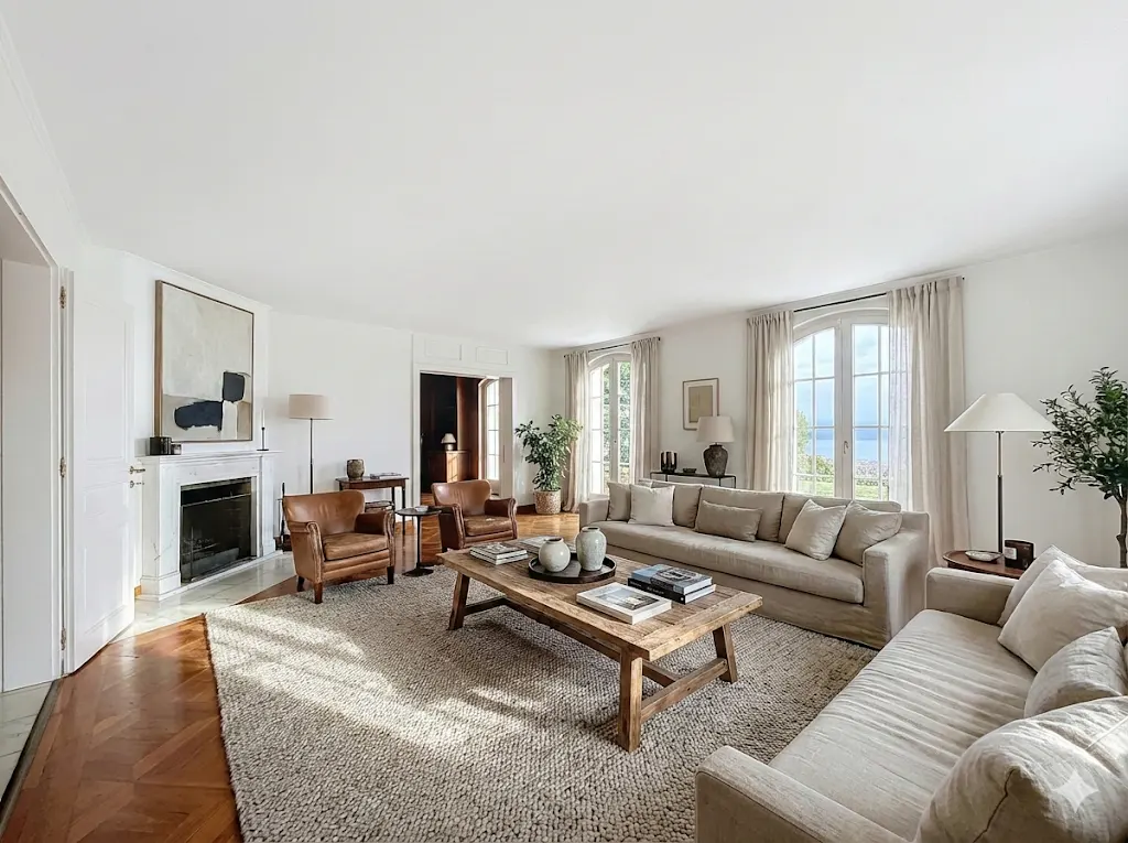Bright living room with white walls, hardwood floors, and a large area rug. Two sofas face each other, with a rustic wood coffee table in between. Fireplace and leather chairs.
