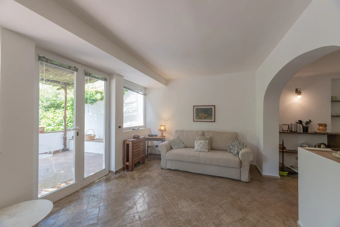 Bright living room with beige sofa, patterned pillows, and tile floor. Glass doors lead to a patio with greenery. Archway to kitchen.
