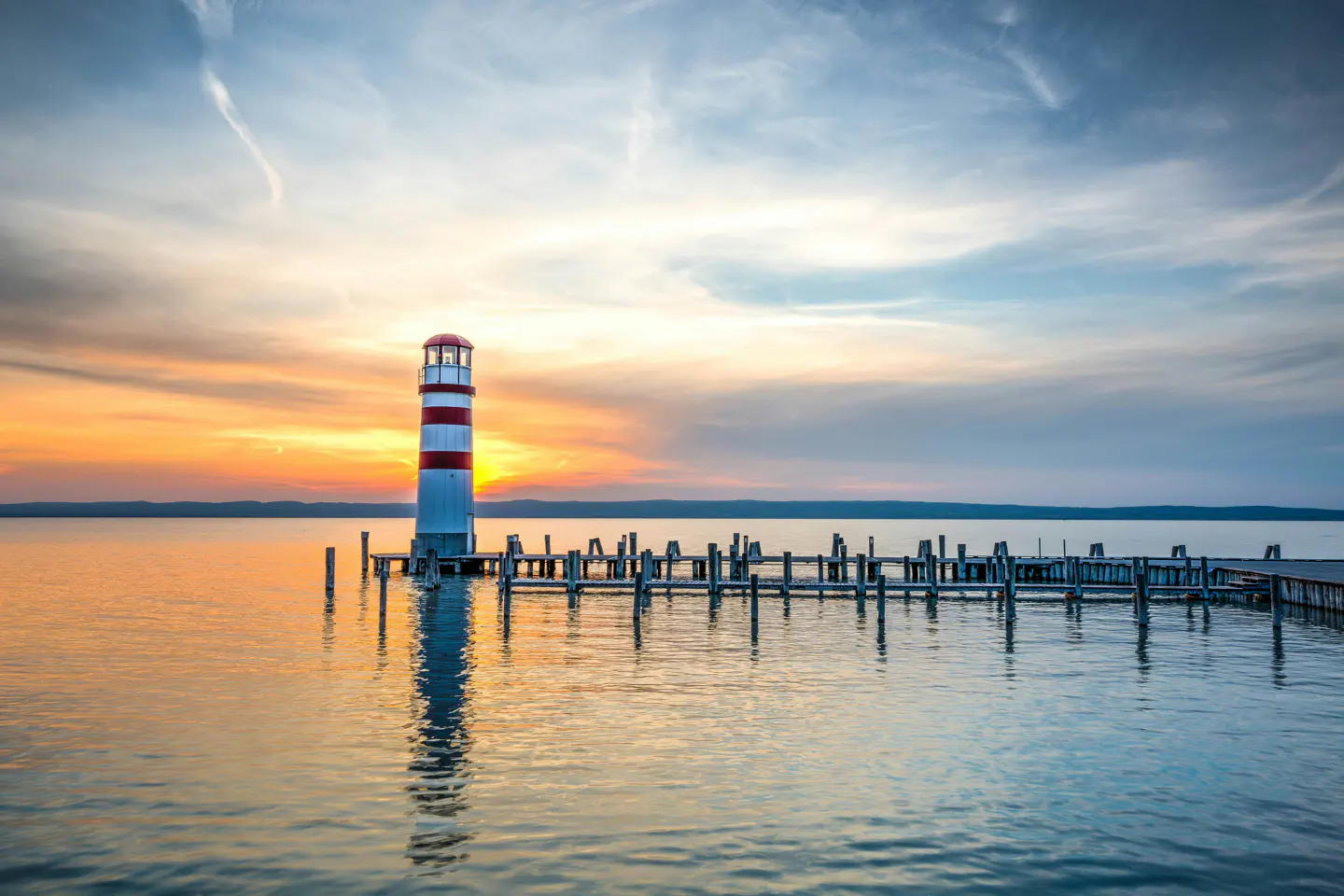 Lighthouse with red and white stripes on a wooden pier at sunset. Water reflects the sky and lighthouse.