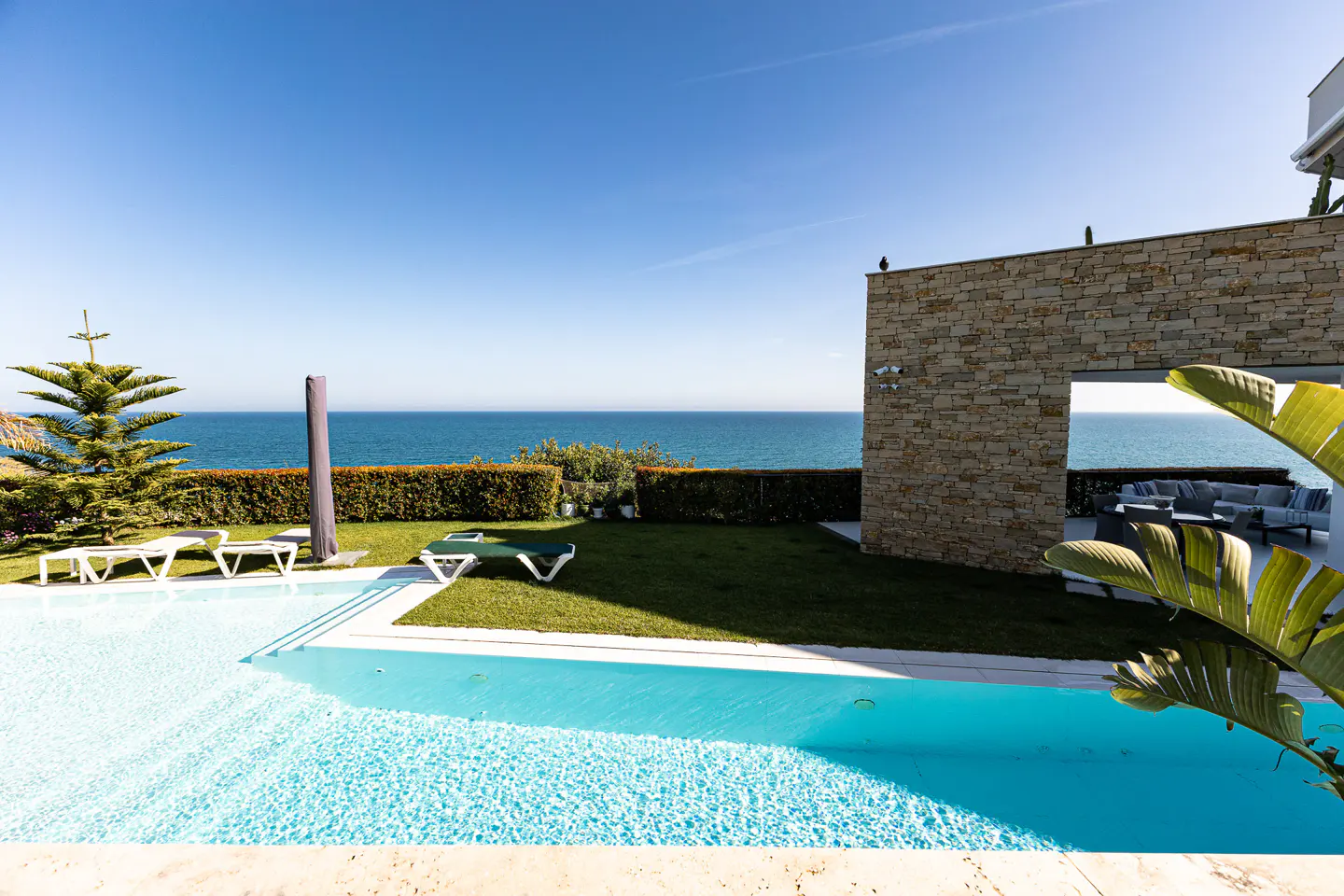 A bright, sunny view of a turquoise pool, green lawn, and the ocean beyond, with lounge chairs and a stone building.