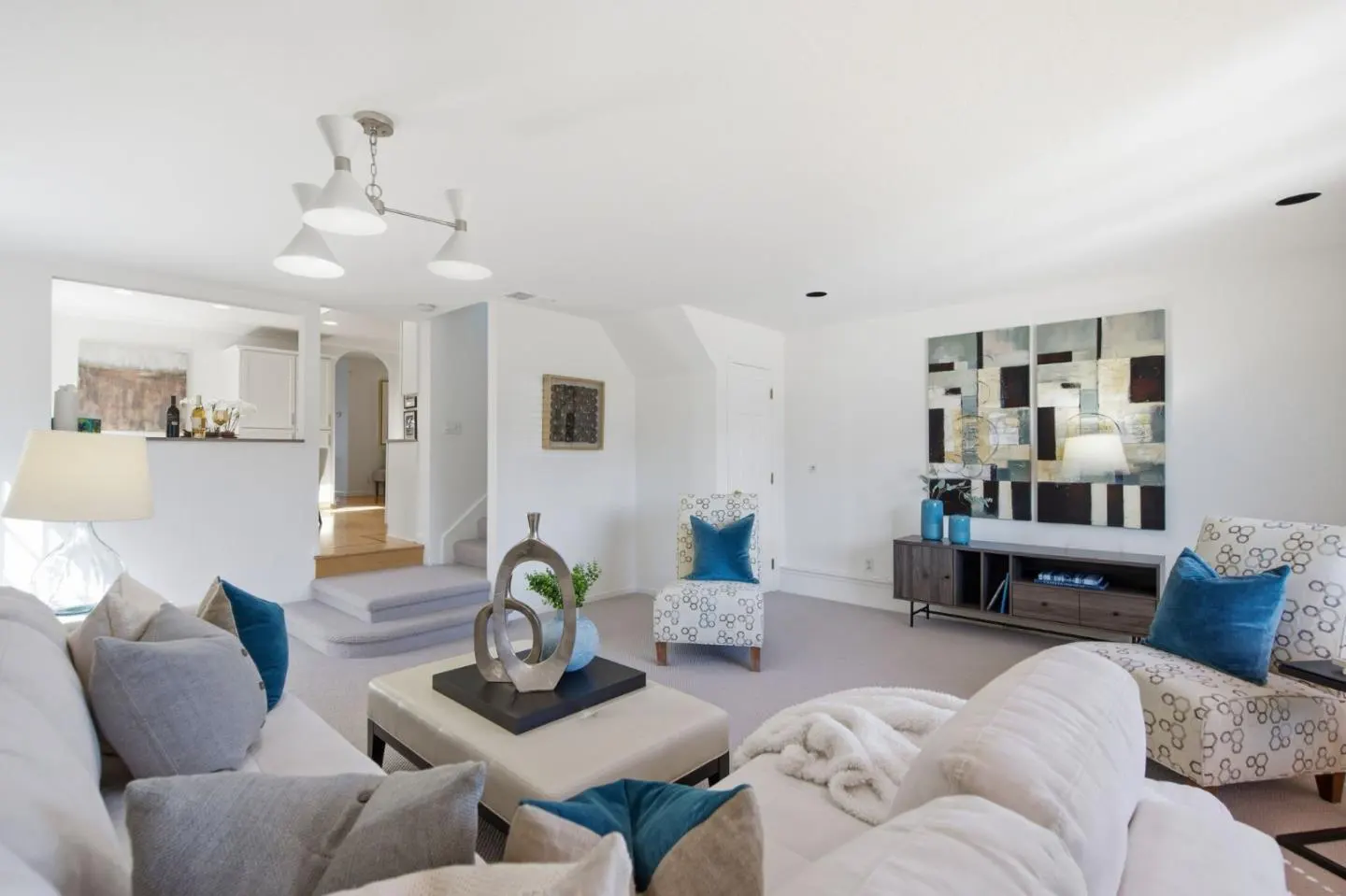 Bright living room with white walls, a white sofa with blue and gray pillows, and patterned armchairs. Abstract art hangs above a wooden console table.