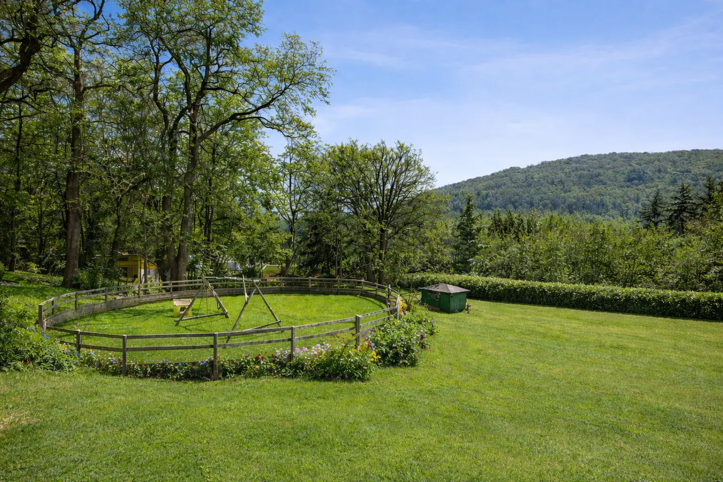 A green lawn with a wooden fence enclosing a swing set, a green shed, trees, and a mountain in the background.