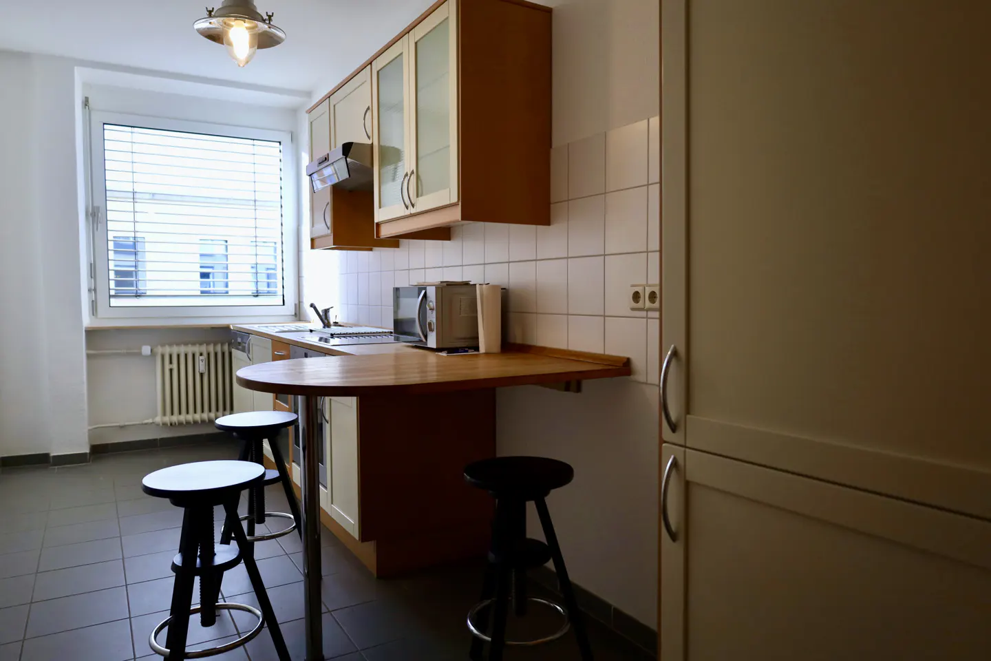 A kitchen with a breakfast bar, stools, and cabinets. A window with blinds is on the left. The floor is tiled.