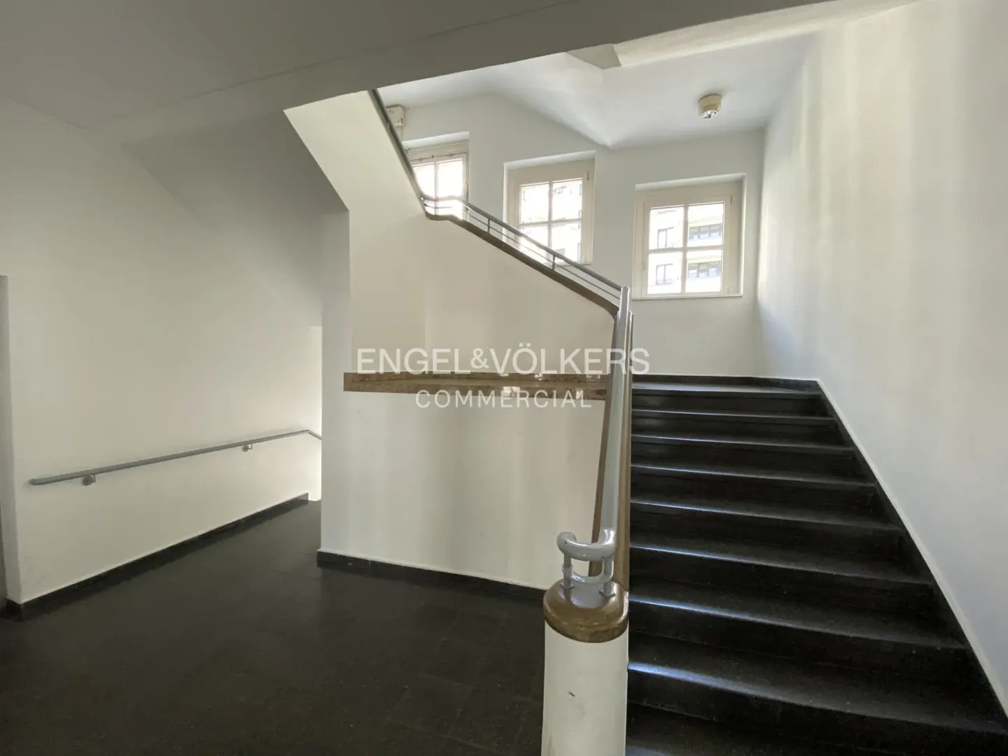 Interior view of a stairwell with black stairs, white walls, and metal handrails. Windows provide natural light. "Engel & Völkers Commercial" logo visible.