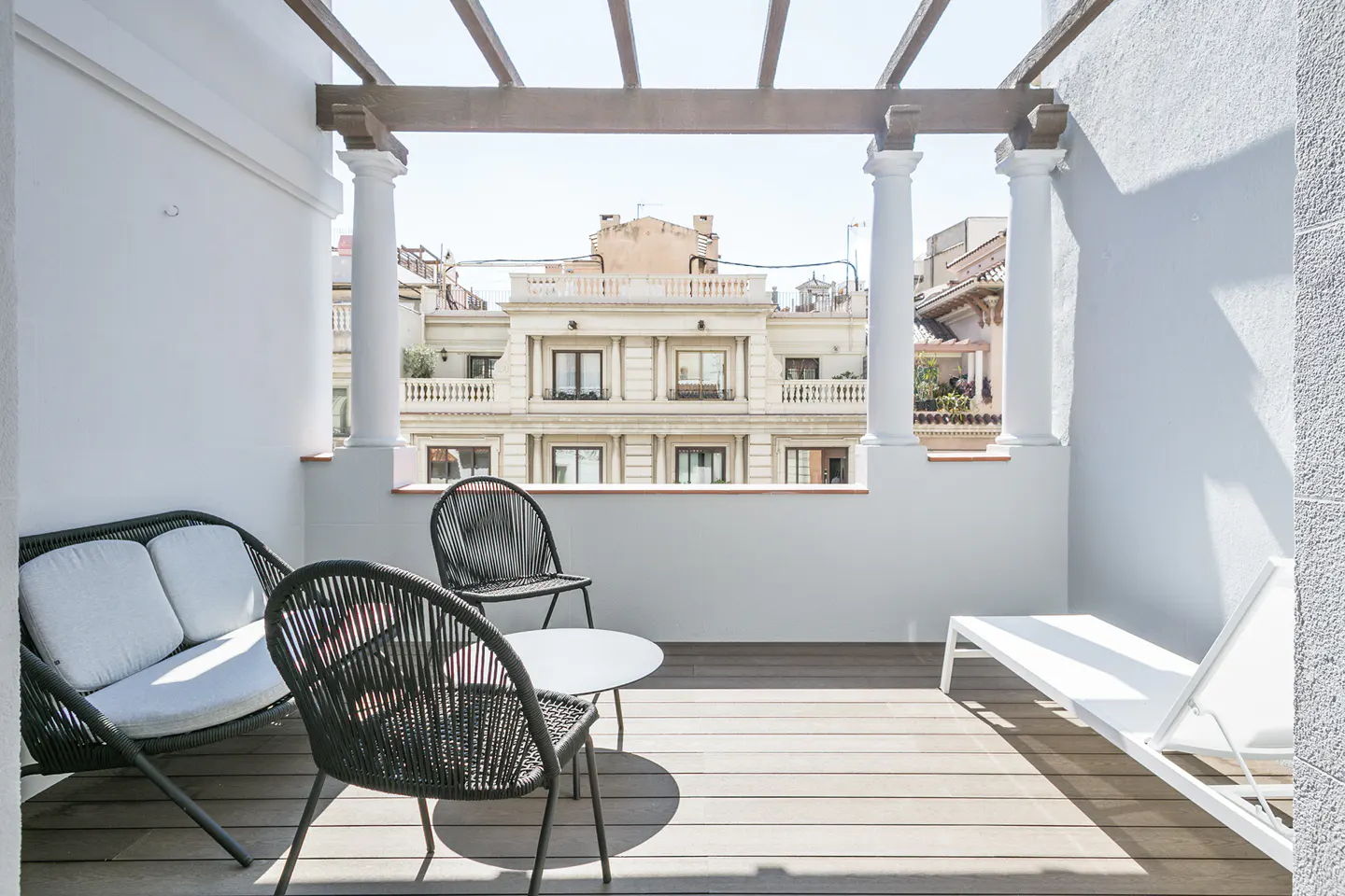 A bright balcony with a wooden floor, white walls, and a view of a building. There is a black sofa, chairs, and a white lounge chair.