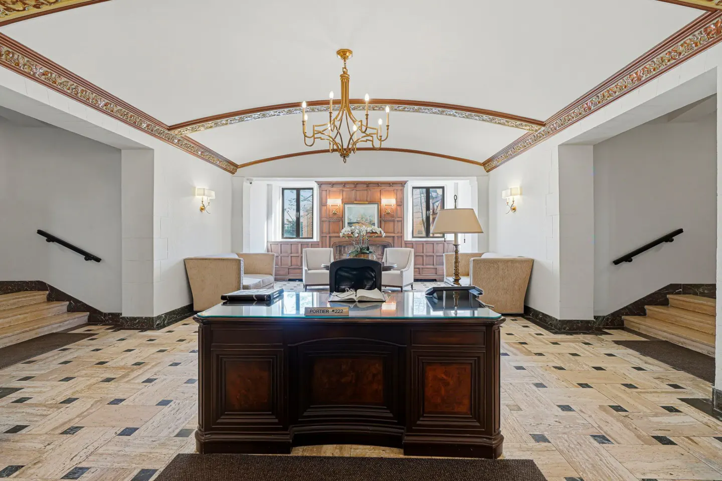 Lobby with a dark wood desk, glass top, and open book. Seating area with chairs, wood paneling, and a gold chandelier.
