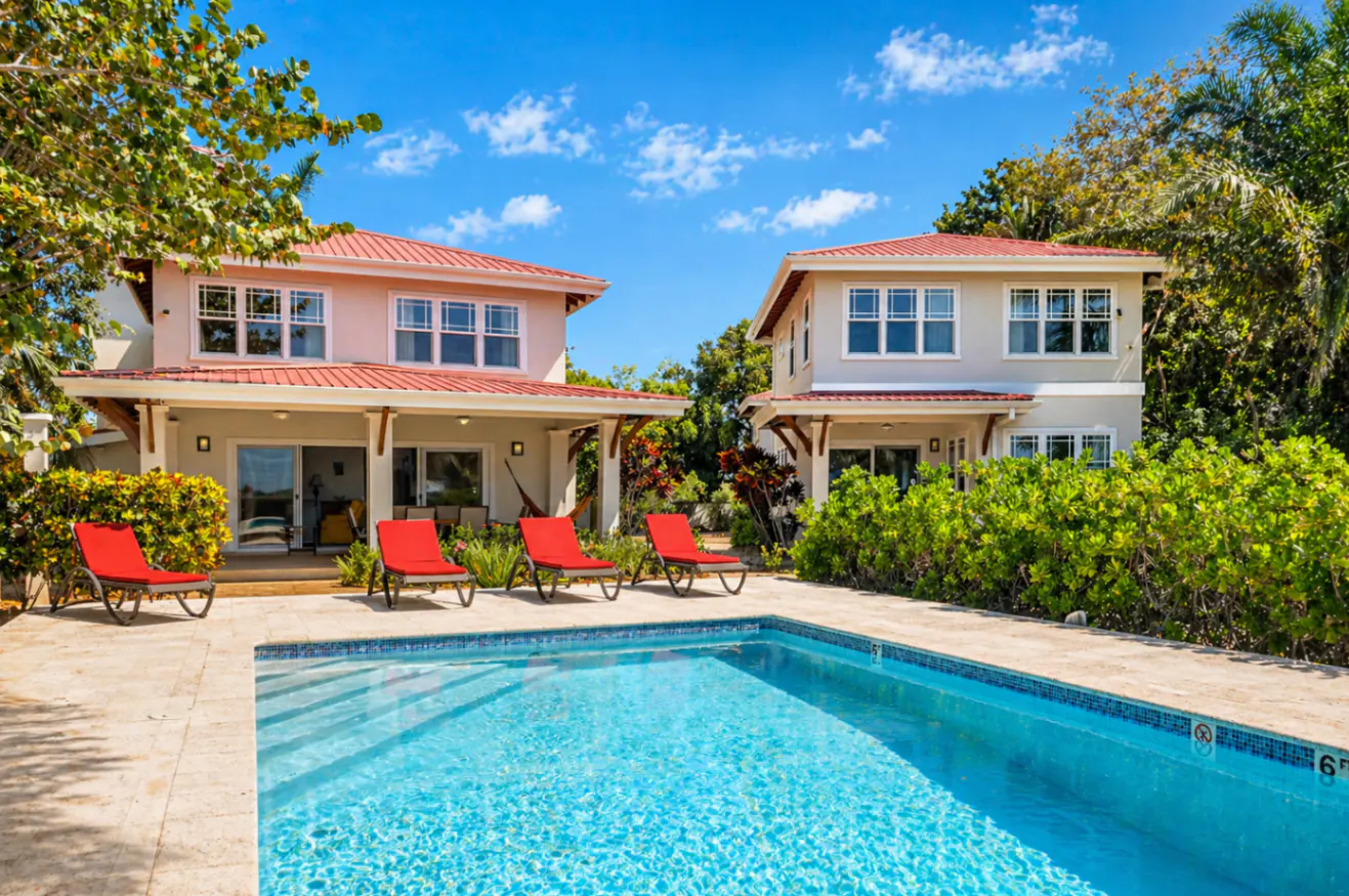 Two-story houses with red roofs flank a blue swimming pool with red lounge chairs on a sunny day.
