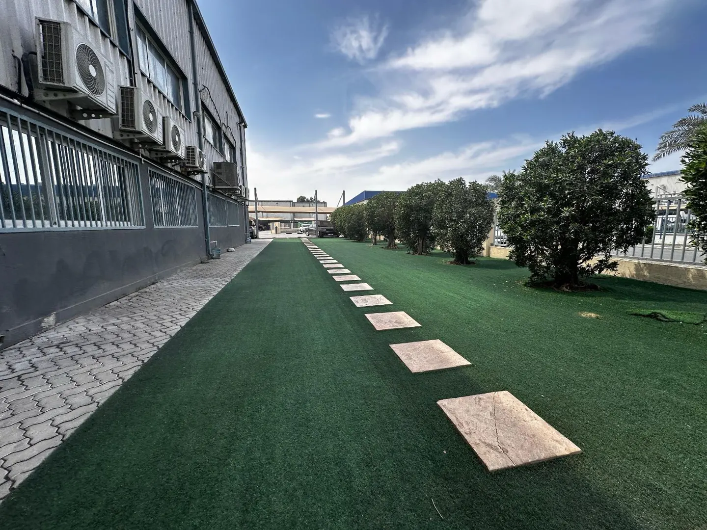 Exterior view of a gray industrial building with AC units, a green lawn, and a stone pathway under a blue sky.