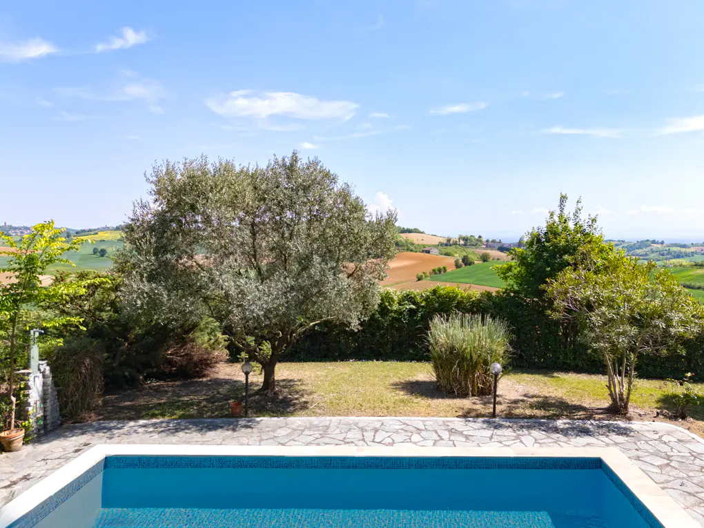 A blue swimming pool with a stone patio overlooks a green lawn, trees, and rolling hills under a blue sky.