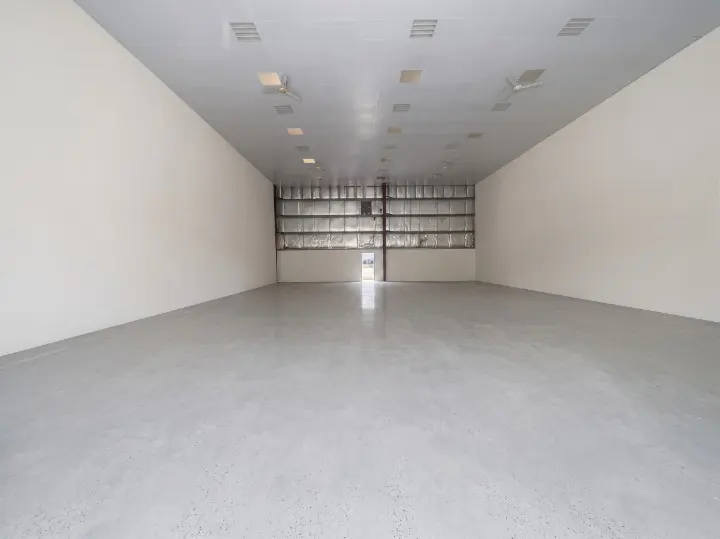Empty commercial space with gray concrete floor, white walls, and a metal ceiling with fluorescent lights. A large metal door is at the back.