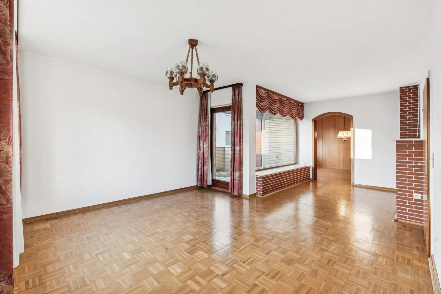Empty living room with parquet wood floors, white walls, a rustic chandelier, and red brick accents near an arched doorway.