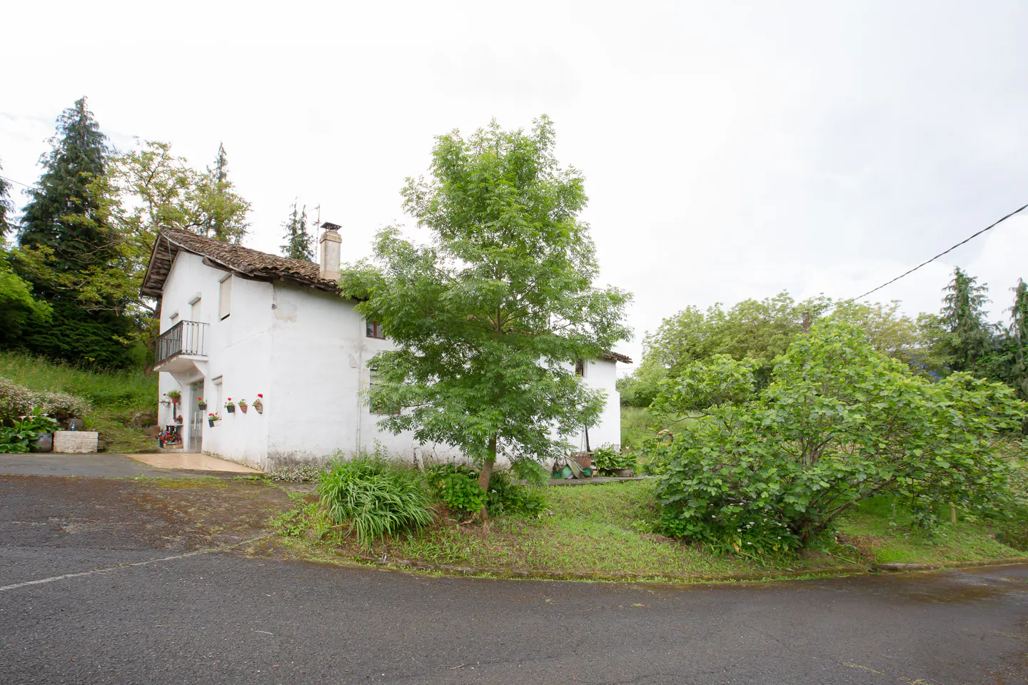 A two-story white house with a brown roof and a small balcony, surrounded by green trees and bushes.