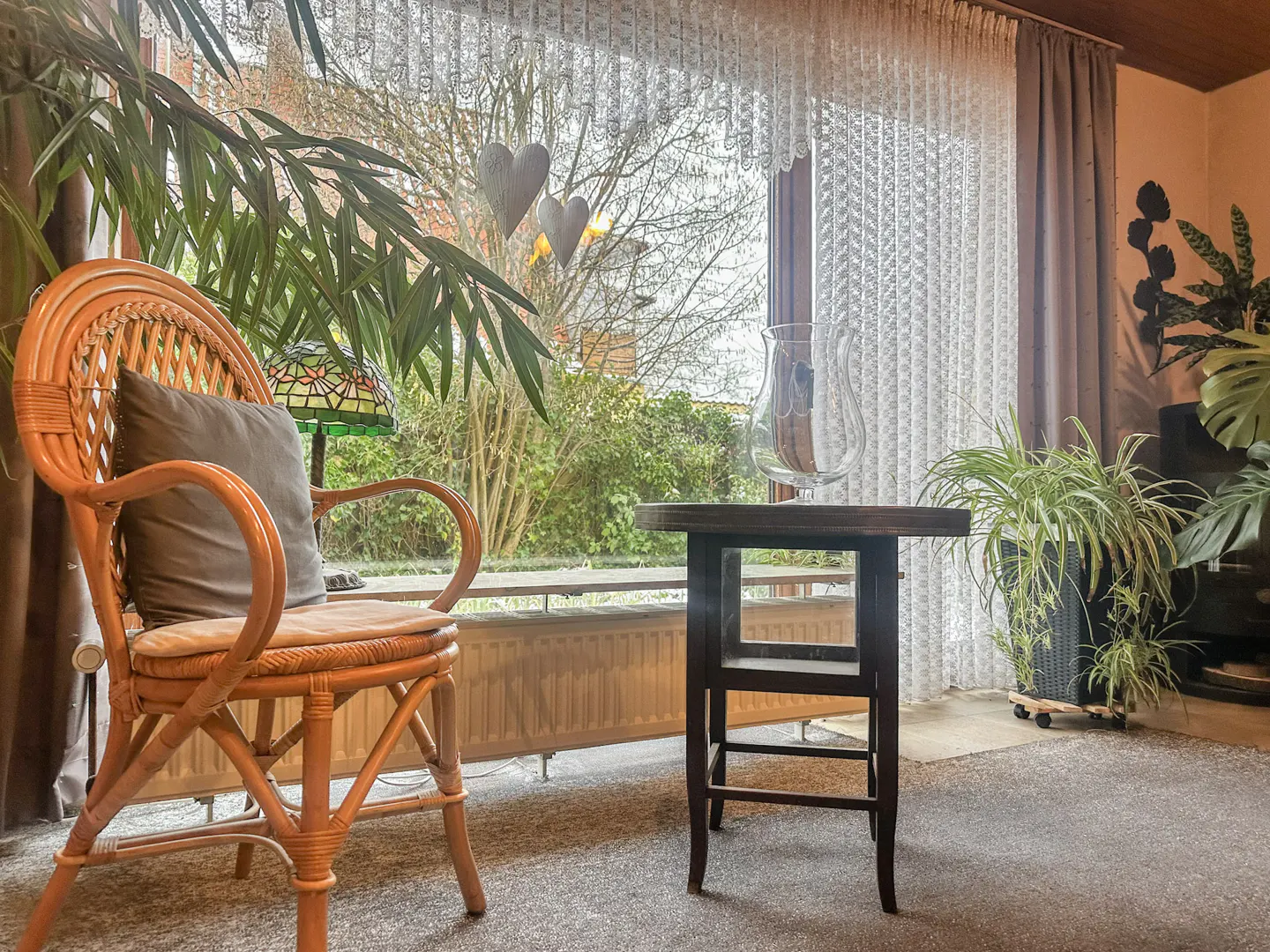 Cozy sunroom with a wicker chair, brown pillow, dark wood table, and large window with lace curtains. Green plants add a touch of nature.