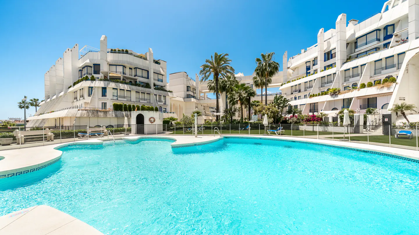 A turquoise pool is surrounded by white buildings and palm trees under a clear blue sky.