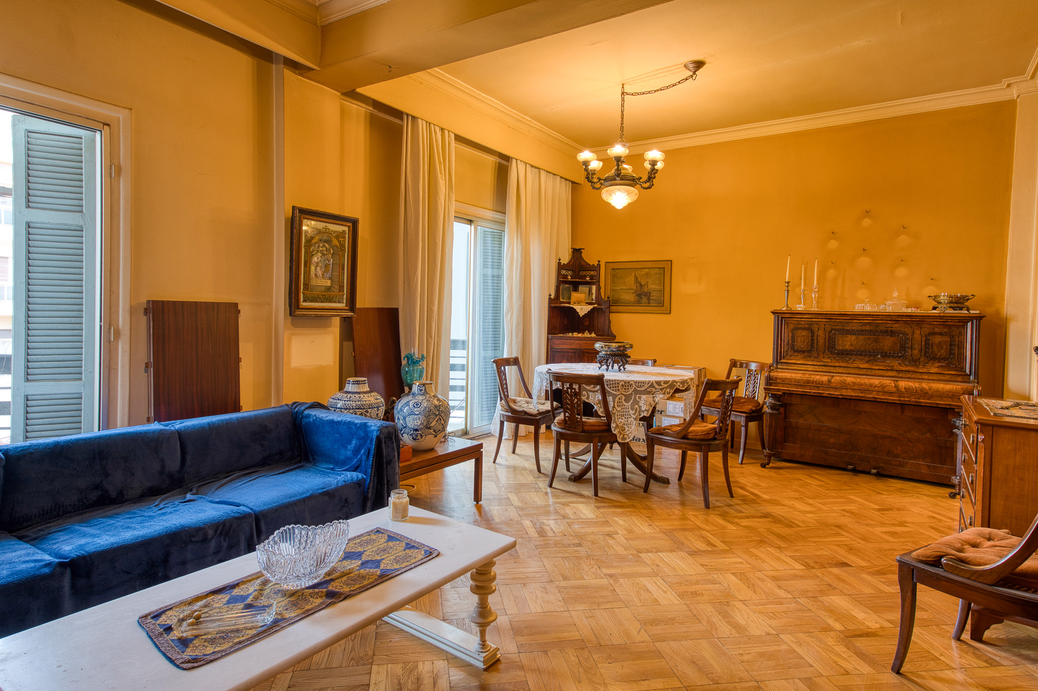 A classic living room with a blue sofa, white coffee table, dining set, and upright piano on parquet wood floors.