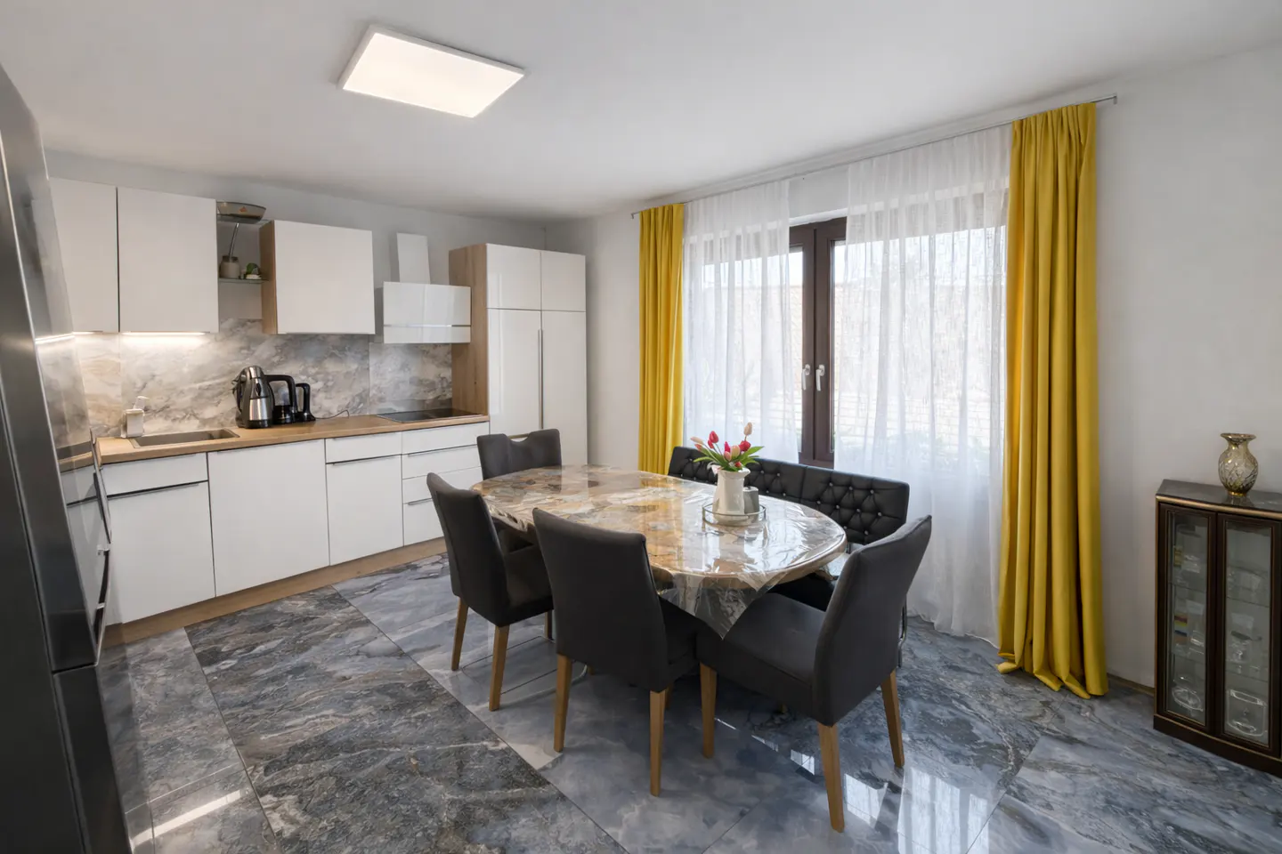 A modern kitchen and dining area with white cabinets, gray marble floors, and yellow curtains. An oval table with gray chairs sits in the center.