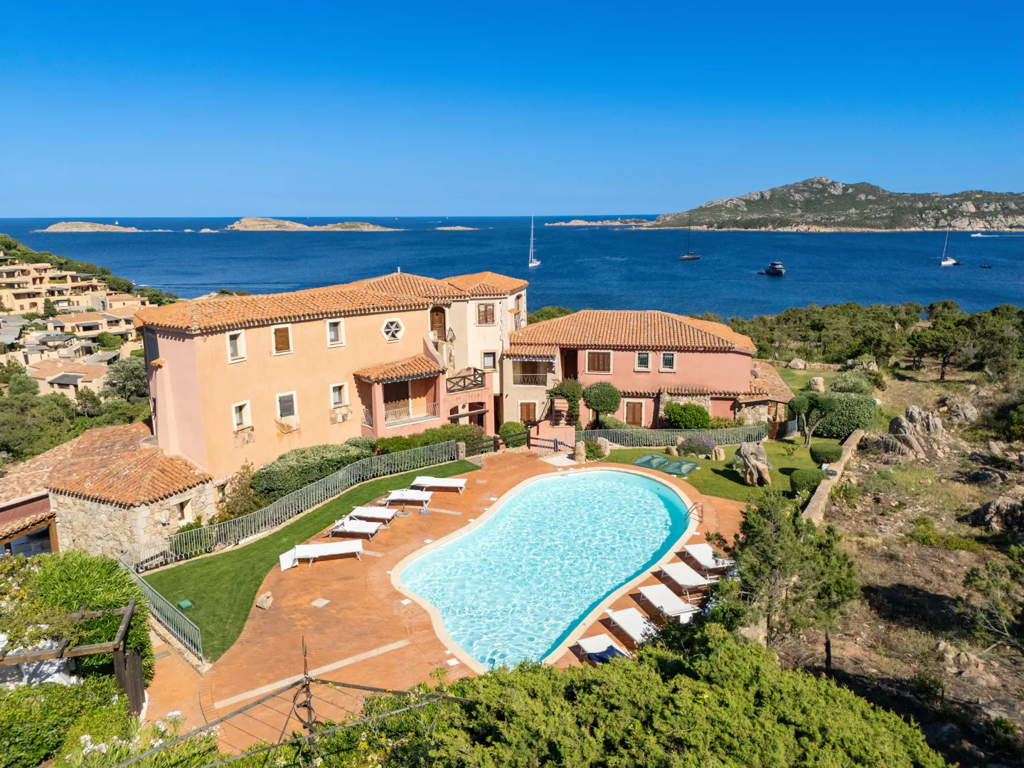 A peach-colored villa with a swimming pool overlooks a blue sea with sailboats under a clear sky.