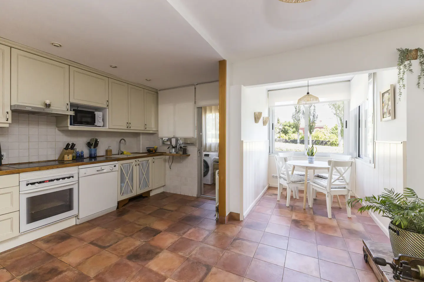 A bright kitchen with terracotta tile flooring, cream cabinets, and a white dining nook with a view.