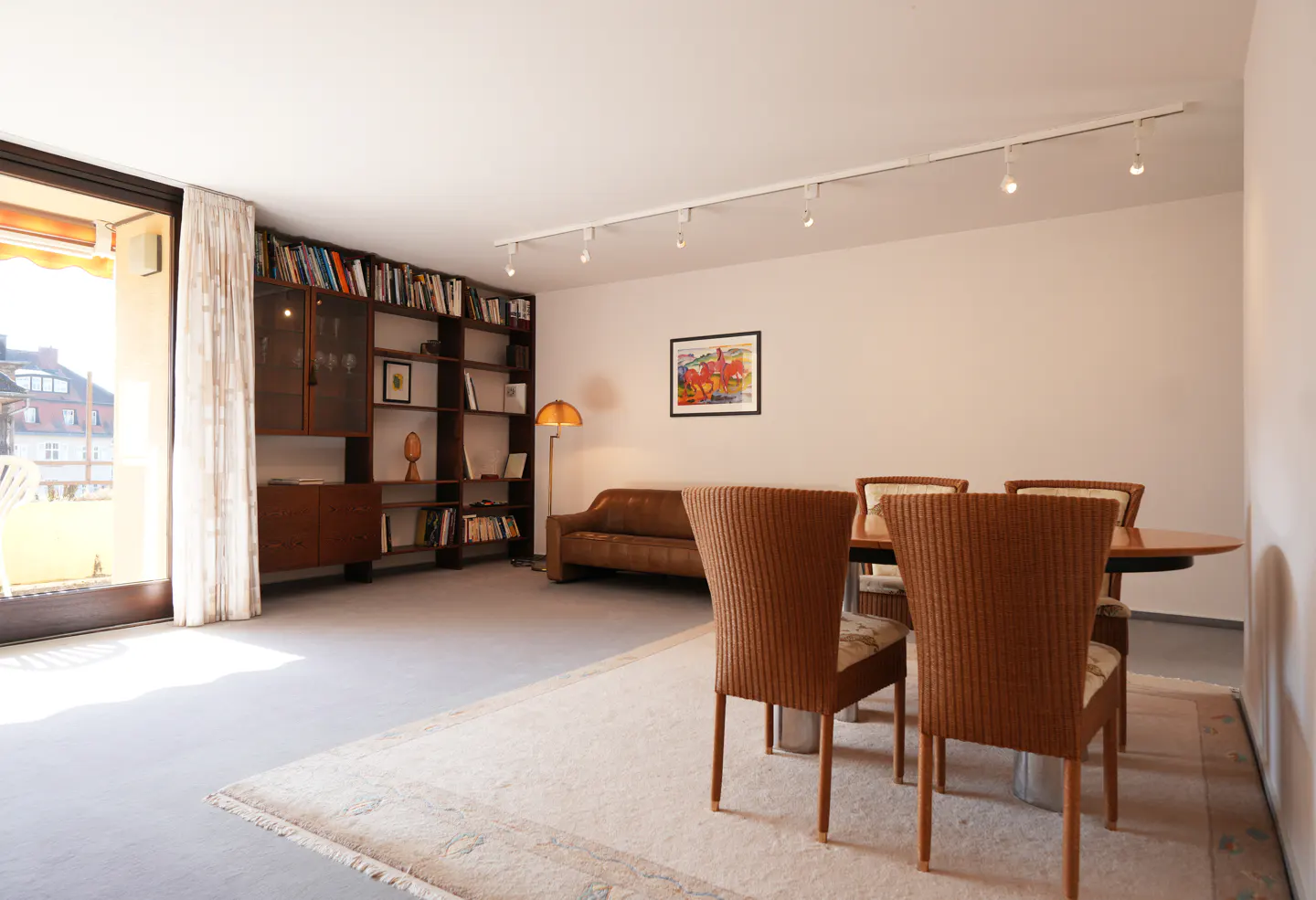 A bright living room with a bookshelf, brown sofa, and a dining table with four wicker chairs. A balcony door lets in natural light.