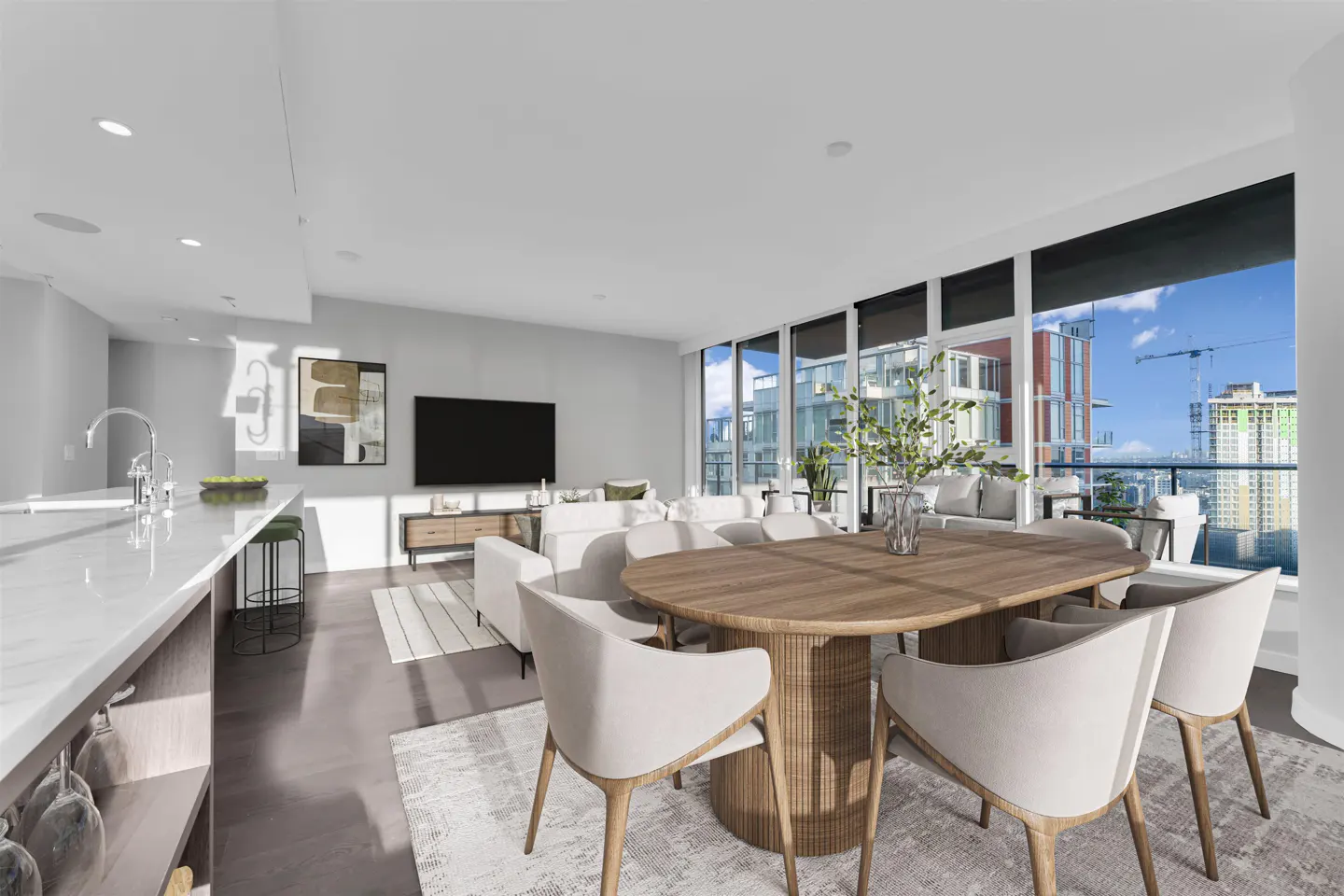 Bright, open-concept condo with a wood dining table, white chairs, and a city view from large windows. Modern kitchen and living area.