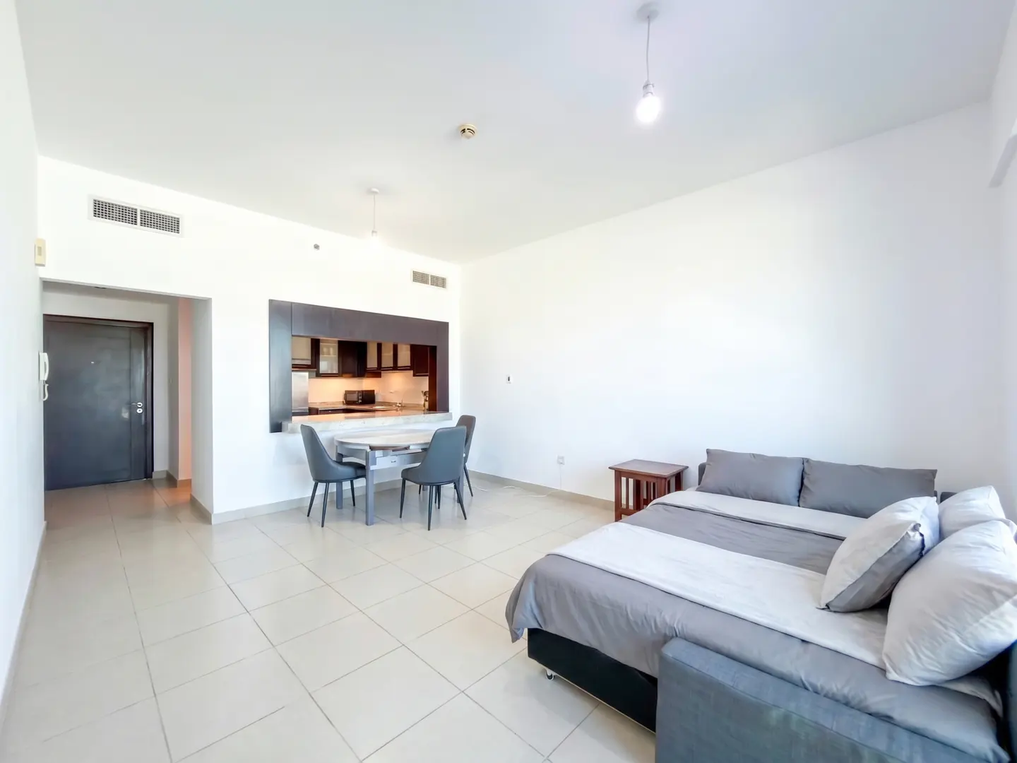 Studio apartment with white walls, tile floors, a grey sofa bed, and a dining area with dark chairs and kitchen cabinets.