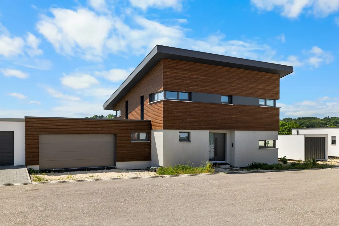 Modern two-story home with wood siding, white stucco, and a flat roof under a bright blue sky with scattered clouds.