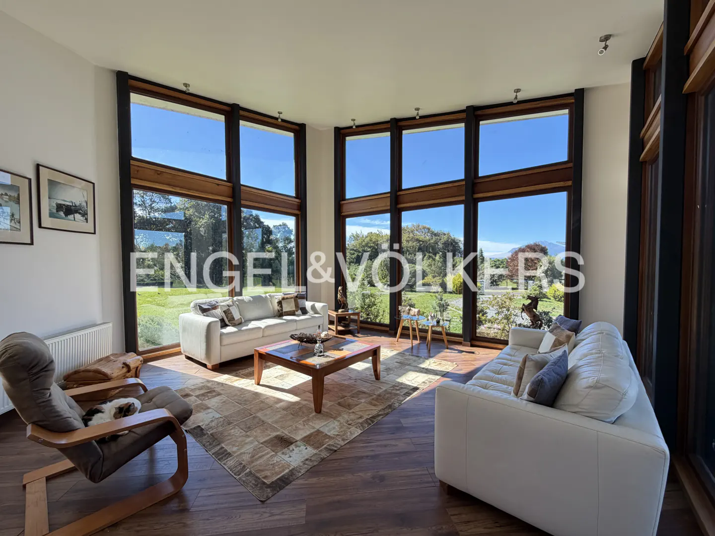Bright living room with large windows, wood floors, and white sofas. A dog sleeps in a chair. Outside is a green lawn and blue sky.