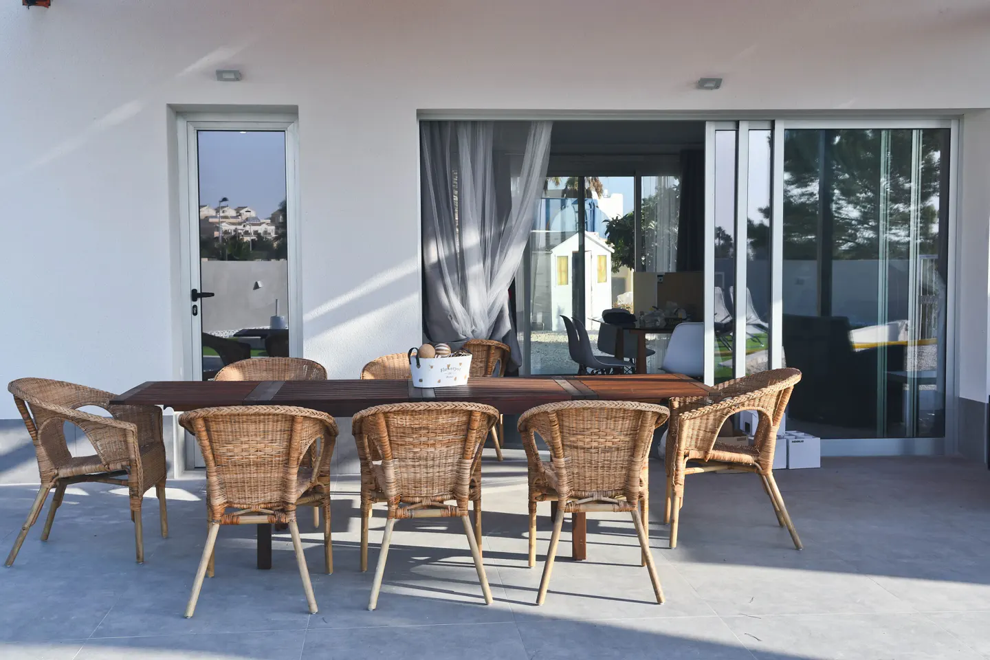 Outdoor patio with a dark wood table and eight wicker chairs on a gray tile floor. Sliding glass doors and a white wall are in the background.