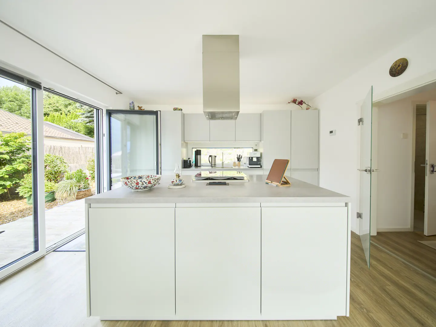 Bright, modern kitchen with white cabinets, island, and stainless steel range hood. Large glass doors open to a lush green garden.
